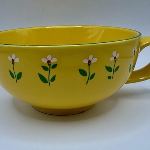 May include: A large, yellow ceramic teacup with a handle. The cup is decorated with white flowers with red centers and green leaves. A thin green line borders the rim of the cup. The cup is on a white background.
