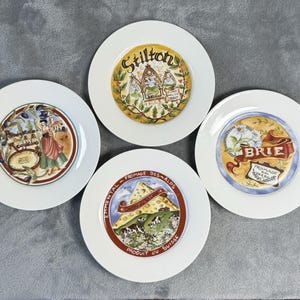 May include: Four white ceramic plates with colorful illustrations. Each plate features a different design: "Stilton" with a castle, "Brie" with flowers, "Parmigiano Reggiano" with a woman, and "Emmental" with a cheese wedge and mountains.