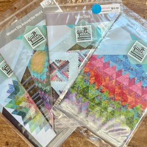 Könnte beinhalten: Vier Quiltmuster in Plastiktüten. Die Muster sind mit "Modern Trip Around the World", "Medallion", "Retro Tiles" und "Rainbow Layer Cake" betitelt. Jedes Muster zeigt ein anderes Quiltdesign und ist mit dem Logo "Quilt Addicts Anonymous" gekennzeichnet.