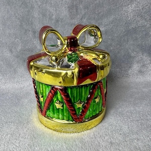 Puede incluir: Una caja de regalo navideña decorativa con borde y lazo dorados. La caja tiene una base verde con cinta roja y estrellas doradas. El lazo es rojo con contorno dorado. Adorno festivo navideño.