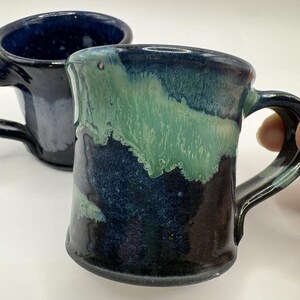 May include: Two ceramic mugs with handles. The front mug has a dark blue base with green and blue glaze. The second mug is dark blue. The mugs are handmade and have a rustic, organic feel. The mugs are suitable for coffee, tea, or other beverages.