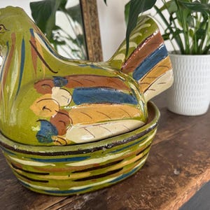 Może przedstawiać: Ręcznie malowany ceramiczny pojemnik w kształcie kury w odcieniach zieleni, żółci, niebieskiego i brązu. Kura siedzi na pasującej podstawie w kształcie kosza. Ten dekoracyjny element jest prawdopodobnie przeznaczony do przechowywania lub ekspozycji.