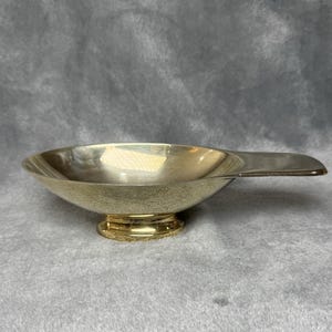 May include: A silver-colored ashtray with a round bowl and a handle. The ashtray has a textured surface and sits on a small, decorative base. The background is a solid gray color.
