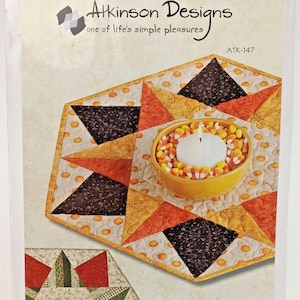 May include: A Happy Holidays tree skirt and table topper pattern. The design shows a quilted, hexagonal table topper with orange, brown, and yellow triangles. A yellow bowl filled with candy corn is in the center. The package includes the text "Atkinson Designs".