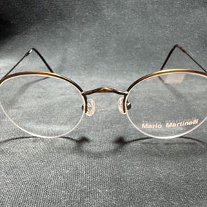 May include: Oval-shaped eyeglasses with a gold-colored rim and dark brown temples. The lenses are clear, and the brand name "Mario Martinelli" is printed on the lower right lens. The glasses have a classic, minimalist design.