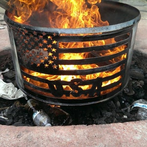 May include: A metal fire pit with a cut-out American flag design and the word "USA" in the center. The fire pit is burning with flames inside.