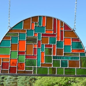 May include: A stained glass window with a geometric design in shades of green, orange, brown, and red. The window is shaped like a half-circle and is hanging from a chain.