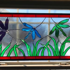 May include: A stained glass window featuring three colorful dragonflies with green, blue, and purple wings. The dragonflies are set against a background of green grass.