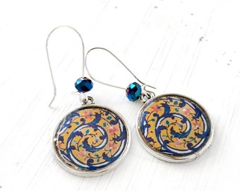 Può includere: Un paio di orecchini color argento con un design circolare. Gli orecchini presentano uno sfondo giallo con motivi floreali blu e rosa. Ogni orecchino ha una perla di cristallo blu e un gancio d'argento.