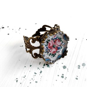 Puede incluir: Un anillo ajustable con una banda de metal en tono bronce antiguo con intrincados filigranas. El engaste circular sostiene un cabujón de vidrio que muestra un patrón floral impreso de rosas rosadas y pequeñas flores azules sobre un fondo blanco.