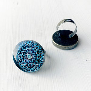 Puede incluir: Dos anillos ajustables con un diseño redondo cubierto de vidrio. El diseño presenta un patrón floral azul y negro. Los anillos tienen una banda de metal plateado y una base de metal oscuro.