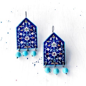 May include: A pair of blue and turquoise earrings with a floral design. The earrings are made of metal and have a geometric shape. The earrings are hanging from a white surface.