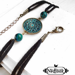 Puede incluir: Un brazalete de cuero negro con un dije de metal redondo con un diseño turquesa y negro. El dije tiene un diseño ornamentado.