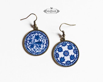 Puede incluir: Un par de pendientes de cerámica azul y blanca con un diseño de pavo real en un pendiente y un diseño floral en el otro. Los pendientes están hechos de metal y tienen un cierre de gancho.