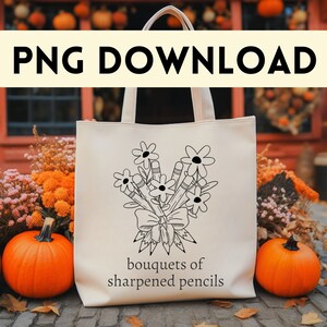 Pode incluir: Uma bolsa tote branca com um desenho de linha preta de um buquê de lápis amarrados com um laço. O texto "bouquets of sharpened pencils" está impresso abaixo do desenho. A bolsa está sentada em uma calçada de paralelepípedos em frente a um prédio com abóboras.