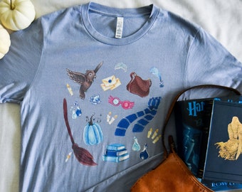 May include: A light blue t-shirt with a watercolor design of various Harry Potter themed objects, including a broom, a book, a scarf, a potion bottle, a pumpkin, and an owl.