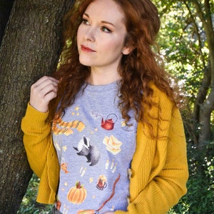 May include: A woman with long, curly red hair is wearing a gray t-shirt with a colorful print of a badger, a teapot, a scarf, and other objects. She is also wearing a yellow cardigan and gray jeans.
