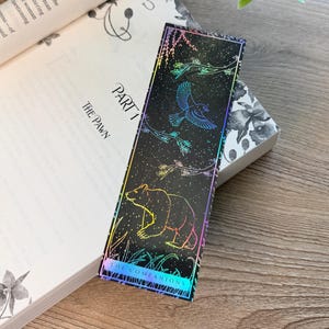 May include: A rectangular bookmark with a black background featuring holographic line art of a bear, a bird, and tree branches. The design includes a thin holographic border and the text "THE COMPANIONS" printed in a clear, sans-serif font at the bottom.