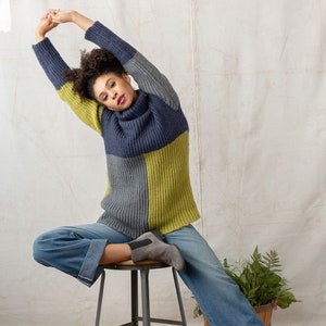 May include: A woman wearing a blue, gray, and yellow color block sweater with a turtleneck. She is sitting on a stool with her arms raised above her head. She is wearing blue jeans and gray ankle boots.