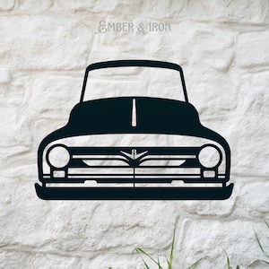 May include: Black metal silhouette of a vintage pickup truck, front view, with a grill and headlights. The truck is on a white brick wall background. The text "EMBER & IRON" is above the truck.