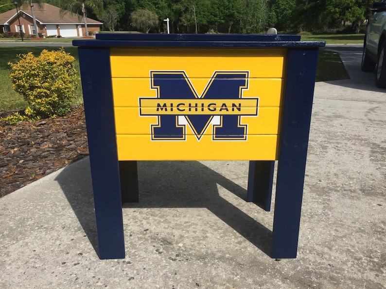 May include: A blue and yellow cooler with the University of Michigan logo and the word "MICHIGAN" printed on the front.