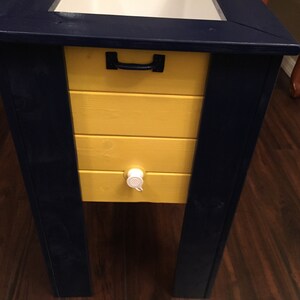 May include: A wooden cooler with a yellow front and dark blue sides. The cooler has a black handle and a white spigot.