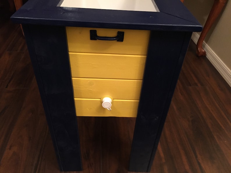 May include: A wooden cooler with a yellow front and dark blue sides. The cooler has a black handle and a white spigot.