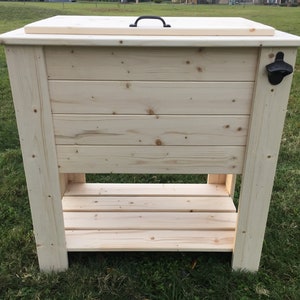 Puede incluir: Una nevera de madera con una tapa con bisagras y un abridor de botellas en el lateral. La nevera tiene un estante debajo para almacenamiento.