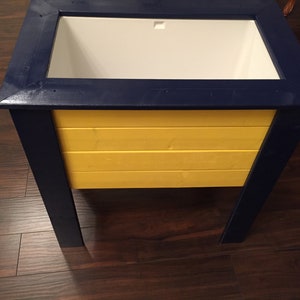 May include: A wooden cooler with a blue frame and a yellow body. The cooler has a white liner and a lid.