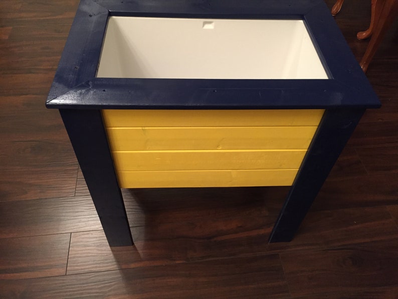 May include: A wooden cooler with a blue frame and a yellow body. The cooler has a white liner and a lid.