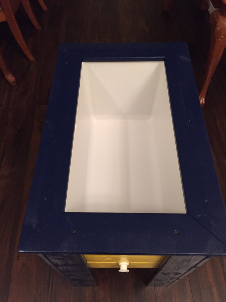 May include: A blue and yellow wooden cooler with a white interior. The cooler has a small drawer with a white knob.