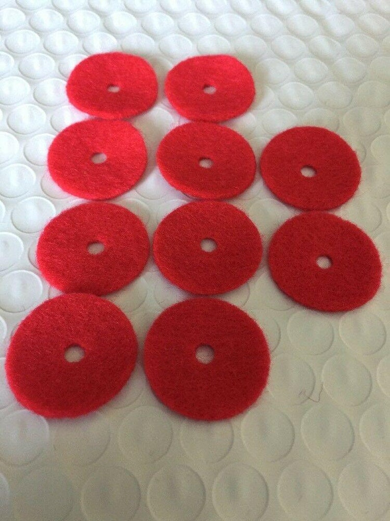 May include: A collection of ten bright red, circular pads with a central hole. The pads are arranged on a white, textured surface. The pads appear to be made of a fibrous material, possibly for cleaning or polishing.