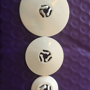 May include: Three white plastic lids of varying sizes, each with a unique central cutout design. The lids are arranged vertically against a textured purple background. The cutouts appear to be for a specific type of container.