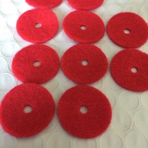 May include: A collection of ten bright red, circular felt pads, each with a central hole. The pads are arranged in a grid-like pattern, showcasing their uniform shape and vibrant color. These pads are likely used for crafts or furniture protection.