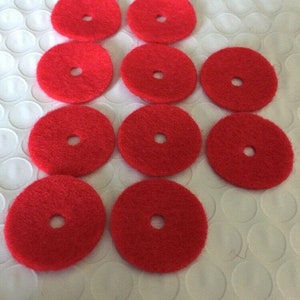 May include: A collection of ten bright red, circular pads with a central hole. The pads are arranged on a white, textured surface. The pads appear to be made of a fibrous material, possibly for cleaning or polishing.