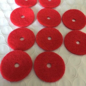 May include: A collection of ten bright red, circular pads with a central hole. The pads are arranged on a white, textured surface. The pads appear to be made of a fibrous material.