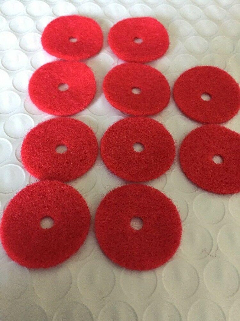 May include: A collection of ten bright red, circular pads with a central hole. The pads are arranged on a white, textured surface. The pads appear to be made of a fibrous material.