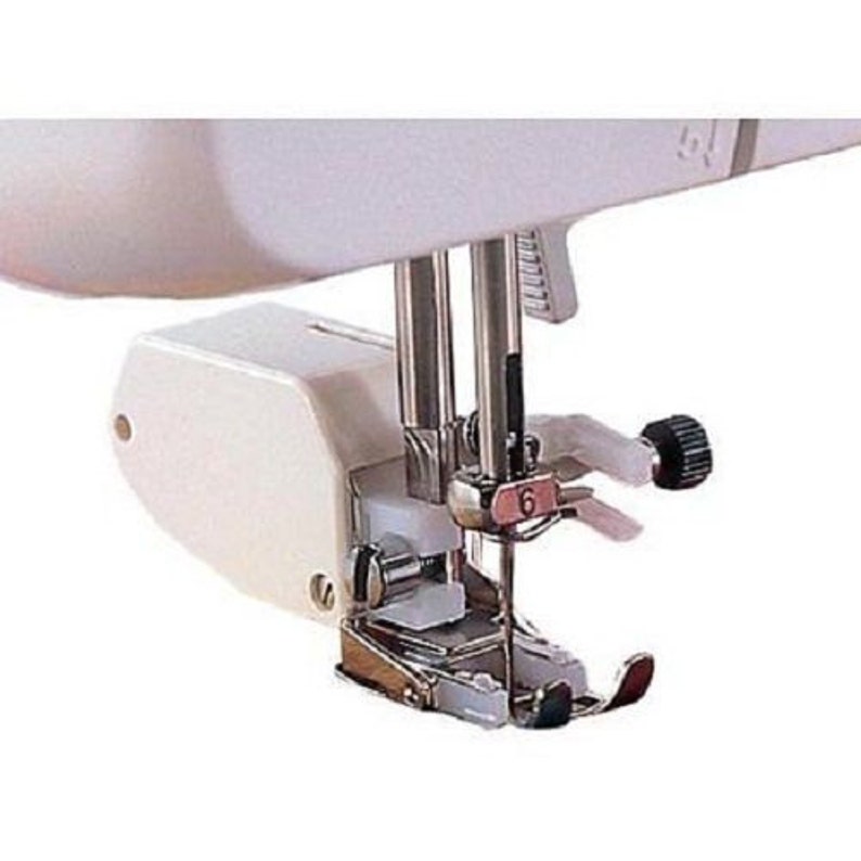 May include: Close-up of a white sewing machine foot with a silver needle and a silver foot lever. The foot is attached to a white sewing machine arm.