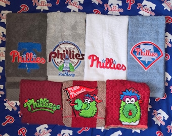 Może przedstawiać: Zestaw 10 haftowanych ręczników z logo Philadelphia Phillies w różnych wzorach. Ręczniki są w różnych kolorach, w tym szarym, białym, niebieskim, czerwonym i zielonym. Ręczniki przedstawiają różne logo, w tym Dzwon Wolności, piłkę baseballową i kreskówkową maskotkę.