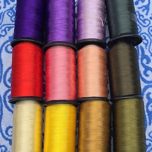 Könnte beinhalten: Zwölf Garnrollen in verschiedenen Farben, darunter Lila, Rot, Rosa, Beige, Gelb, Gold, Grün und Schwarz. Die Rollen sind in drei Reihen zu vier angeordnet.
