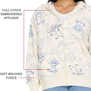 May include: Cream-colored zip-up hoodie with a blue embroidered Stitch design. The hoodie features a full stitch embroidered applique and soft brushed fleece. The design includes Stitch characters, stars, and text. The hoodie has a hood and a zipper closure.