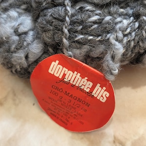 May include: A ball of grey and white speckled yarn with a red tag. The tag reads "dorothée bis" and "CRÔ-MAGNON 100 g - 3 1/2 oz WOOL". The yarn is thick and appears to be made of wool, suitable for knitting or crocheting projects.