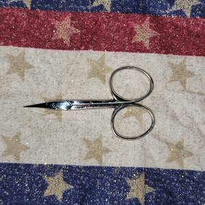 May include: A pair of small, silver-coloured cuticle scissors with a pointed tip. The scissors are lying on a white fabric with gold stars.