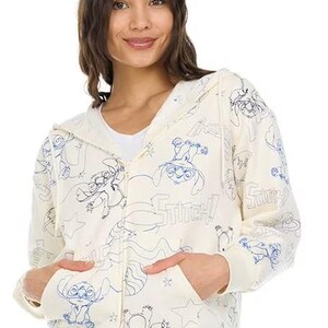 May include: Cream-colored zip-up hoodie featuring blue line art of the character Stitch from Lilo & Stitch. The hoodie has a hood, pockets, and the word "Stitch" printed throughout the design. The model is wearing blue jeans.