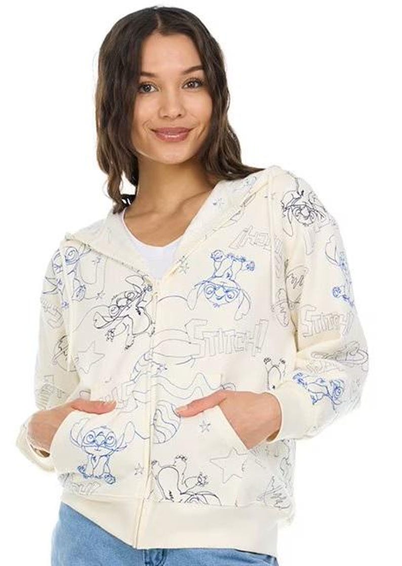 May include: Cream-colored zip-up hoodie featuring blue line art of the character Stitch from Lilo & Stitch. The hoodie has a hood, pockets, and the word "Stitch" printed throughout the design. The model is wearing blue jeans.