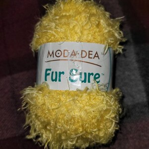 Pode incluir: Uma bola de fio amarelo chamada "Fur Sure" da Moda Dea. O fio é felpudo e tem uma textura felpuda.