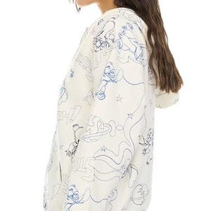 May include: Cream-colored zip-up hoodie with a blue cartoon character outline pattern. The hoodie features a drawstring hood, ribbed cuffs, and a ribbed hem. The design includes stars, planets, and other cartoon elements. The model is wearing blue jeans.