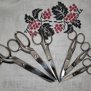 May include: A set of six silver scissors of varying sizes, arranged on a white linen surface with a black and red embroidered design.