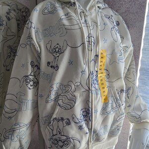 May include: Cream-colored zip-up hoodie featuring a repeating blue outline pattern of the character Stitch, planets, and stars. The hoodie has a drawstring hood, front pockets, and a yellow tag with the size "M".