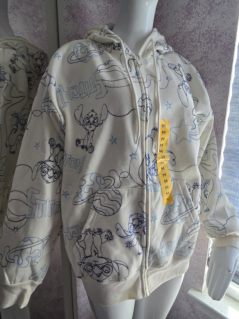 May include: Cream-colored zip-up hoodie featuring a repeating blue outline pattern of the character Stitch, planets, and stars. The hoodie has a drawstring hood, front pockets, and a yellow tag with the size "M".